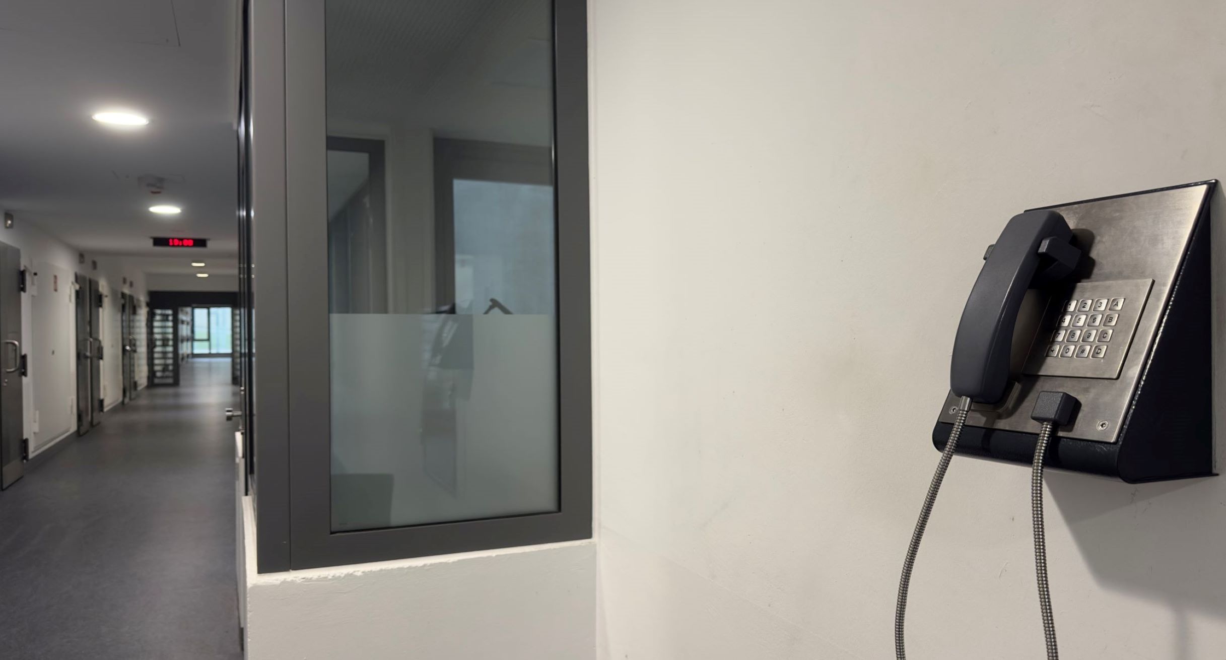 Ein Telefon hängt an der Wand, dahinter ist ein Gang innerhalb einer Justizvollzugsanstalt zu sehen.